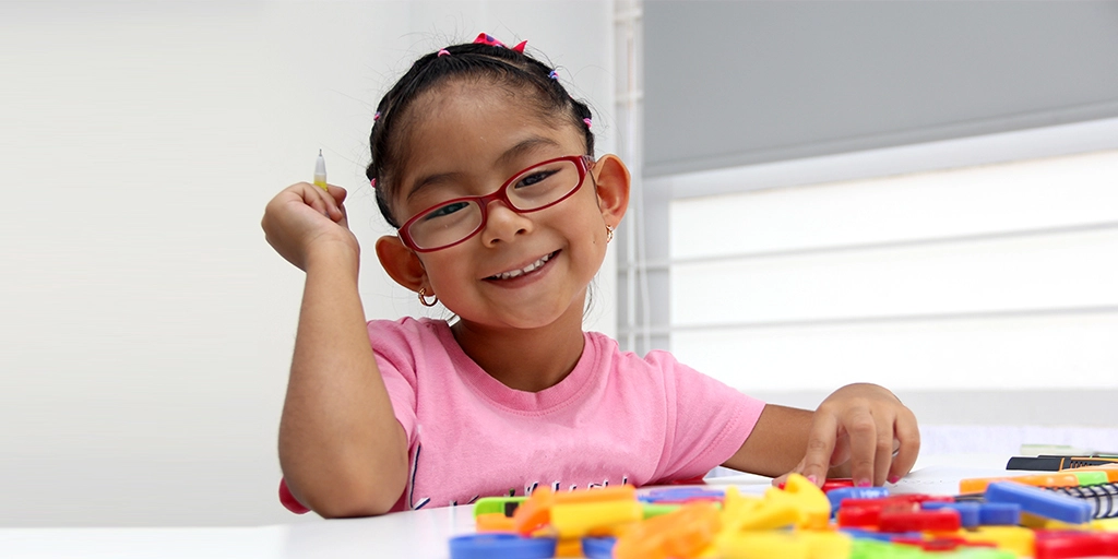 A young girl with autism wearing glasses, smiling, and engaging in a playful educational activity with colorful blocks at a table.